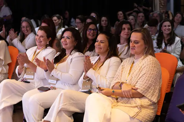 Participantes de branco sorriem e aplaudem na plateia do MulherEHS