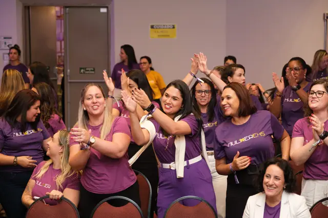 Participantes de camiseta roxa comemorando de braços para o alto
